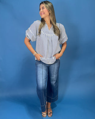 A woman modeling a short-sleeve button-up blouse with vertical blue and white stripes, paired with dark wash jeans and metallic sandals, standing against a solid blue studio backdrop.