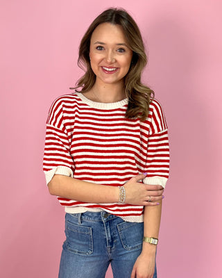 Red and white striped knit short sleeve sweater with a relaxed fit and ribbed trim, paired with high-rise wide leg jeans and pointed metallic flats, styled against a pink background.