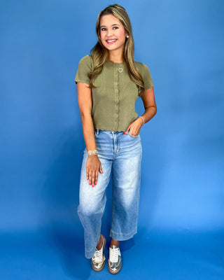 A woman modeling a short-sleeve olive green ribbed top with a button placket, paired with light-wash blue cropped jeans and metallic silver sneakers, set against a solid blue studio backdrop.