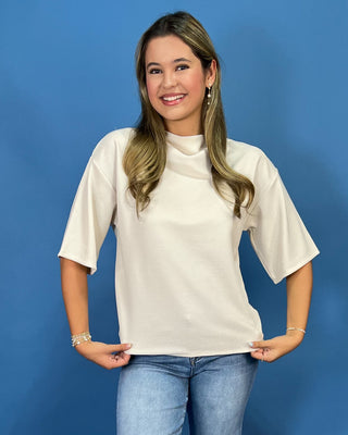 A woman modeling a cream-colored short-sleeve mock neck top with relaxed fit, paired with blue jeans, standing against a solid blue background.