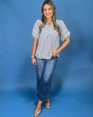 A woman modeling a white short-sleeve blouse with thin blue vertical stripes, paired with blue jeans and metallic sandals, standing against a solid blue studio backdrop.