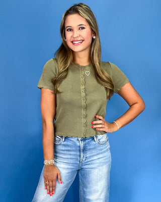 A woman posing in a short-sleeve olive green knit top with a button-down front and light wash blue jeans, set against a solid blue studio background.