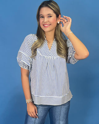 A woman modeling a blue and white vertically striped short-sleeve blouse with puff sleeves and a V-neckline, paired with blue jeans, against a solid blue studio background.