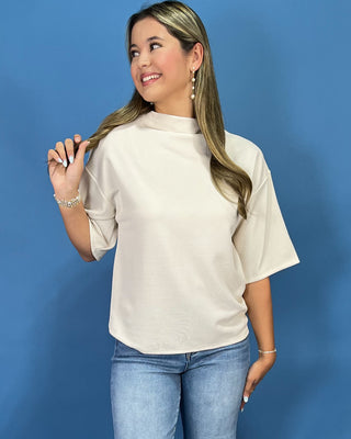 A woman modeling a cream-colored short-sleeve mock neck top with blue jeans, standing against a solid blue studio background.
