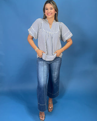 A woman modeling a short-sleeve striped blouse with blue jeans and metallic sandals, standing against a solid blue studio backdrop.