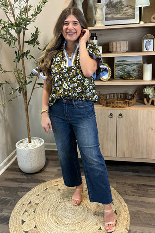 Woman in a floral blouse and jeans standing in a room with decor elements.