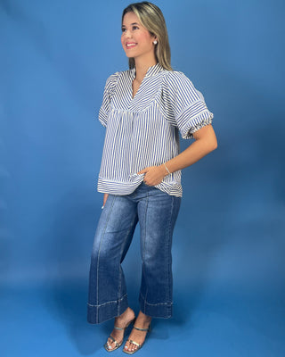 A woman modeling a short-sleeve button-up blouse with vertical navy and white stripes, paired with wide-leg blue jeans and metallic sandals, set against a solid blue studio backdrop.