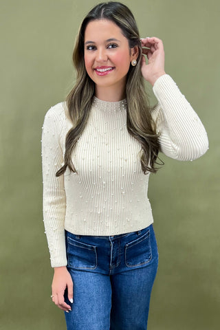 Woman wearing a cream ribbed sweater and blue jeans against a green background