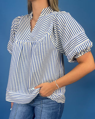 A woman wearing a blue and white vertically striped short-sleeve blouse with puff sleeves and a V-neckline, paired with blue jeans against a solid blue background.