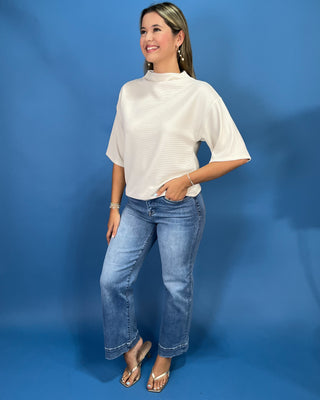 A woman modeling a cream-colored short-sleeve mock neck top with blue jeans and metallic sandals, standing against a solid blue studio backdrop.