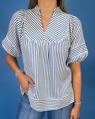 A woman wearing a blue and white vertically striped short-sleeve blouse with puff sleeves and a split V-neckline, paired with blue jeans against a solid blue background.