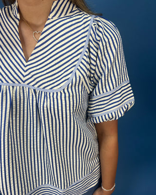 A close-up of a person wearing a short-sleeve blouse with vertical blue and off-white stripes, a V-neckline, and gathered puff sleeves, set against a solid dark blue background.