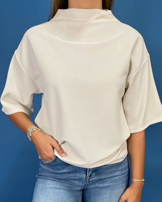 A woman wearing a cream-colored, textured short-sleeve mock neck top and blue jeans, standing against a solid blue background.