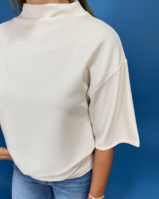 A person wearing a ribbed cream mock-neck top with elbow-length sleeves, paired with blue jeans, set against a solid blue background.