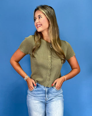 A woman wearing a short-sleeve olive green knit top with a button-down front and light-wash blue jeans, standing against a solid blue background.