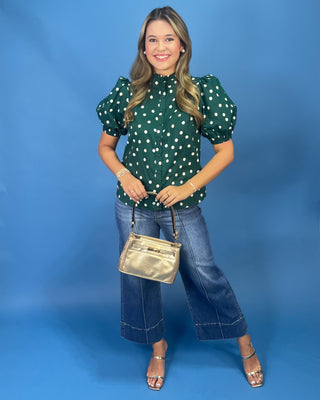 A woman modeling a dark green short-sleeve blouse with white polka dots, paired with wide-leg blue jeans, a metallic gold handbag, and clear-strap high-heeled sandals, set against a solid blue studio backdrop.