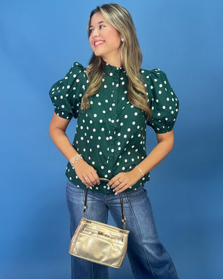 A woman modeling a dark green puff-sleeve blouse with white polka dots, paired with blue jeans and a metallic gold crossbody bag, set against a solid blue studio background.