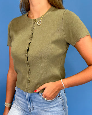 A woman wearing a short-sleeve ribbed knit top in olive green with scalloped edges and a button-front closure, paired with light blue jeans against a solid blue background.