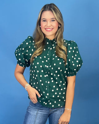 A woman modeling a dark green short-sleeve blouse with white polka dots and puff sleeves, paired with blue jeans, against a solid blue studio background.