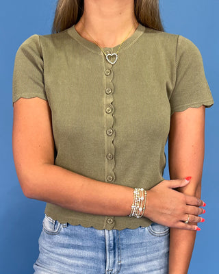 A woman wearing a short-sleeve ribbed knit top in olive green with a scalloped hem and button-front detail, paired with light blue jeans against a solid blue background.