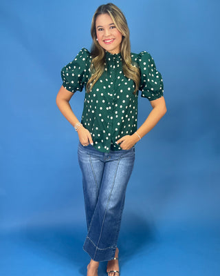 A woman modeling a dark green short-sleeve blouse with white polka dots and puff sleeves, paired with blue jeans, against a solid blue studio background.