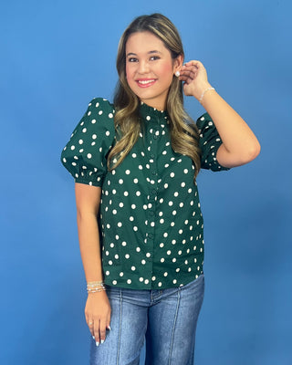 A woman modeling a dark green short-sleeve blouse with white polka dots, paired with blue jeans, against a solid blue studio background.