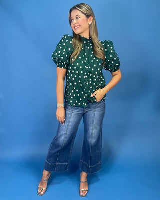 A woman modeling a dark green puff-sleeve blouse with white polka dots, paired with wide-leg blue jeans and metallic sandals, standing against a solid blue studio backdrop.