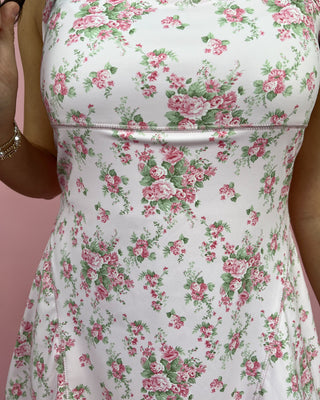 Smiling woman standing against a pink backdrop wearing a white floral mini dress with thin straps and a ruffled hem, styled with metallic gold sneakers.