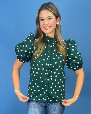 A young woman with blonde hair is wearing a dark green short-sleeve blouse with white polka dots and puff sleeves, paired with blue jeans, against a solid blue background.