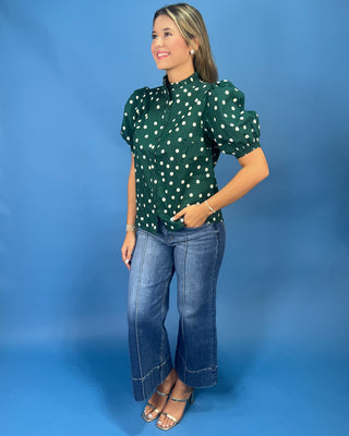A woman modeling a dark green short-sleeve blouse with white polka dots and puffed shoulders, paired with blue jeans and metallic sandals, set against a solid blue studio background.