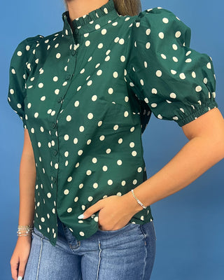 A woman wearing a dark green short-sleeve blouse with white polka dots, featuring puffed short sleeves and a high gathered neckline, paired with blue jeans against a solid blue background.
