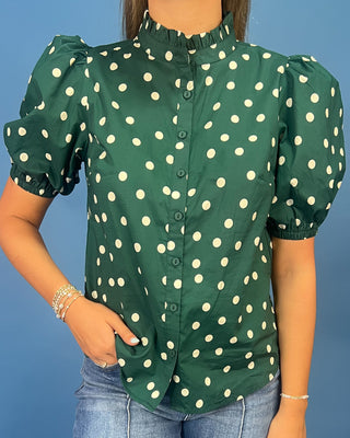 A woman wearing a dark green short-sleeve blouse with white polka dots, featuring a ruffled high collar and puffed sleeves, paired with blue jeans against a solid blue background.