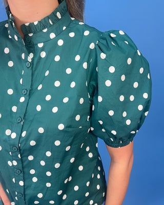 A close-up of a person wearing a teal green short-sleeve blouse with white polka dots, featuring a ruffled high collar and puff sleeves, set against a solid blue background.