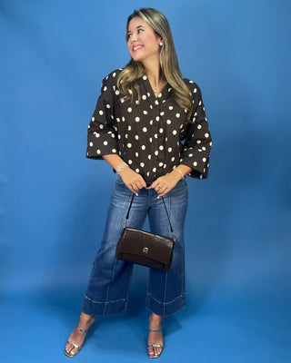A woman modeling a black and white polka dot blouse with elbow-length sleeves, paired with wide-leg blue jeans, a dark brown handbag, and metallic sandals, set against a solid blue studio backdrop.