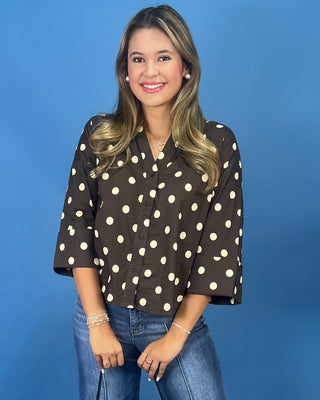 A woman modeling a dark brown button-up blouse with large white polka dots, paired with blue jeans, set against a solid blue studio background.