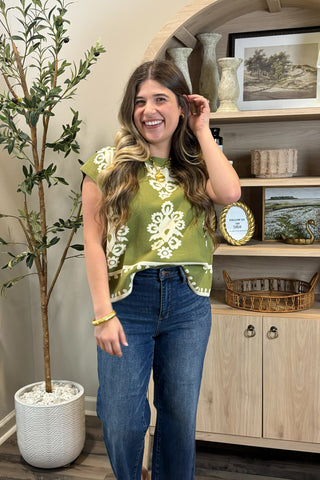 Woman wearing a green top with floral patterns and blue jeans, standing in a room with a plant and wooden cabinet.