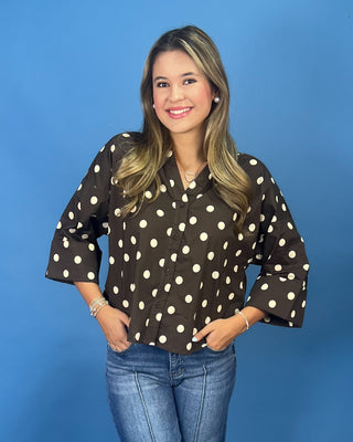 A woman modeling a brown button-up blouse with white polka dots and three-quarter bell sleeves, paired with blue jeans, set against a solid blue background.