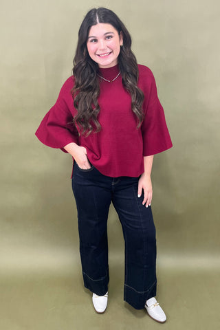 Woman wearing a red sweater and dark jeans against a beige background