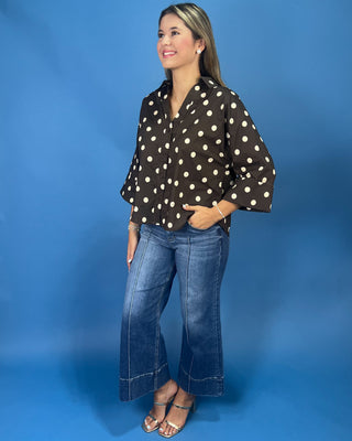 A woman modeling a brown long-sleeve blouse with white polka dots, paired with blue jeans and metallic sandals, standing against a solid blue studio backdrop.