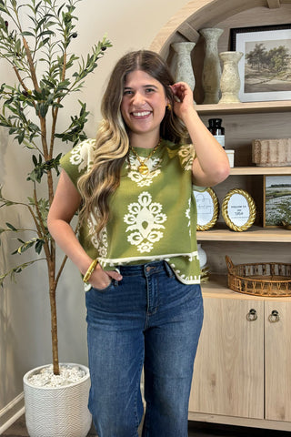 Woman wearing a green patterned top and blue jeans standing in a room with decor elements.