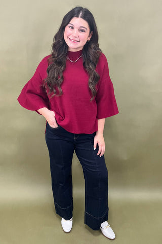 Woman wearing a red top and dark jeans against a beige background