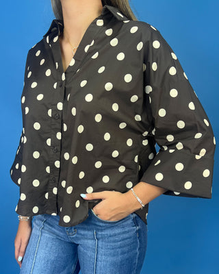 A woman wearing a black button-up blouse with large white polka dots, paired with blue jeans, standing against a solid blue background.