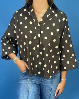 A woman wearing a dark brown button-up blouse with large cream polka dots and blue jeans, standing against a solid blue background.