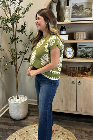 Woman wearing a green and white patterned top in a room with a plant and wooden cabinet.