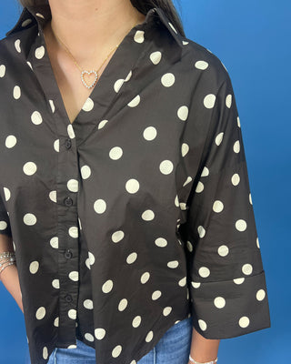 A woman wearing a black button-up blouse with large white polka dots, paired with blue jeans and a gold heart necklace, set against a solid blue background.