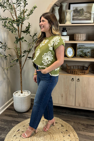 Woman wearing a green floral top and blue jeans standing in a room with a plant and wooden cabinet.