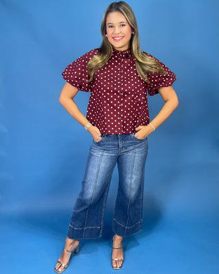 A woman modeling a burgundy short-sleeve blouse with white polka dots, paired with blue wide-leg jeans and silver sandals, standing against a solid blue studio backdrop.