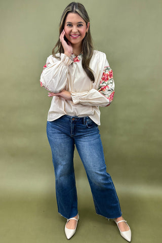Woman wearing a cream blouse with floral sleeves and blue jeans on a green background