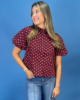 A woman wearing a burgundy short-sleeve button-up blouse with white polka dots and puff sleeves, paired with blue jeans, standing against a solid blue background.