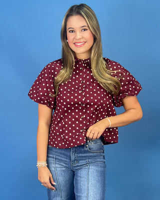 A young woman with blonde hair wearing a burgundy short-sleeve blouse with white polka dots and blue jeans, standing against a solid blue background.